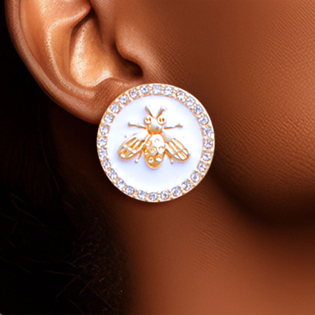Gold bee earrings with white center worn on an ear against a brown background